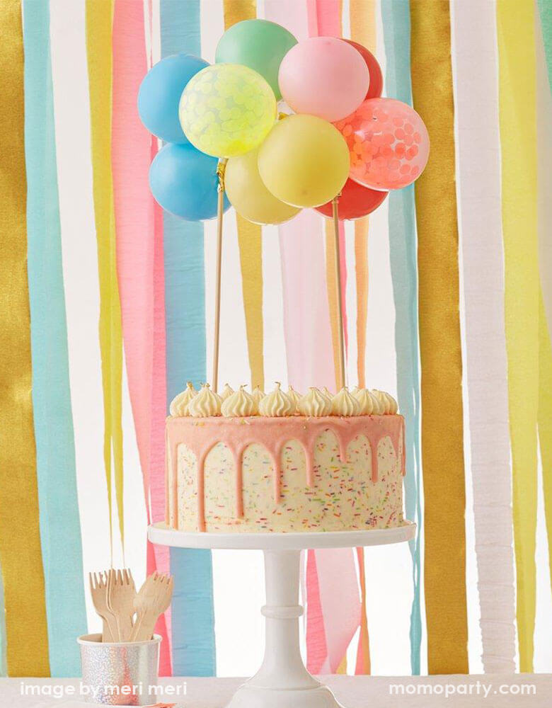 A rainbow themed birthday cake topped with Meri Meri's Rainbow Balloon Cake Topper Kit with mini multicolored latex balloons and confetti balloons in front of a backdrop made with colorful tissue streamers as the curtain 
