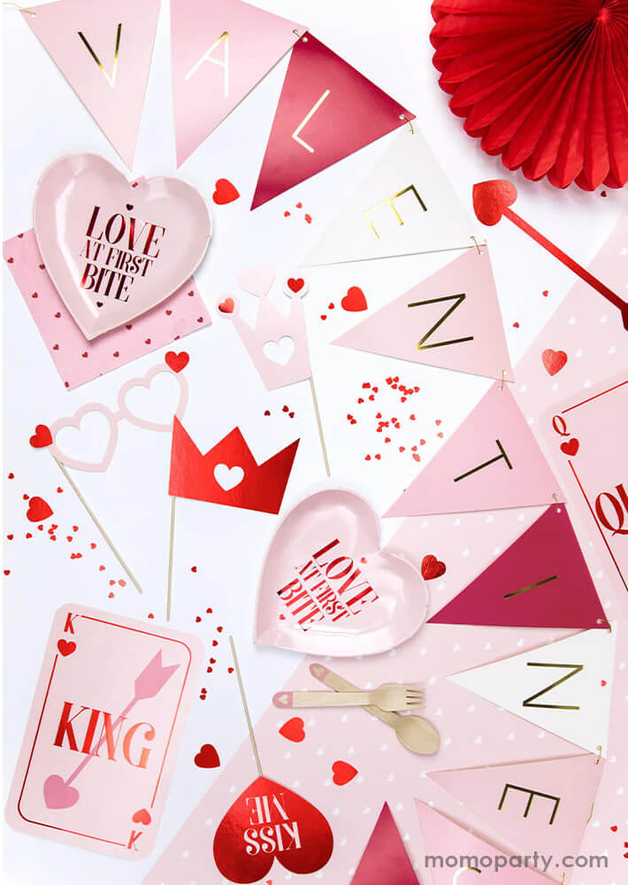 A sweet red and pink Valentine's Day table decoration featuring Party Deco's Valentine's party banner in red and pink hue, fun Valentine's Day photo booth props including heart shaped glasses and "kiss me" heart shaped sign, and Party Deco's "Love at first bite" pink heart shaped plates and pink napkins in heart pattern, plus Party Deco's metallic red heart shaped confetti spread out the party table, it makes a sweet and fun Valentine's Day and Galentine's Day celebration!