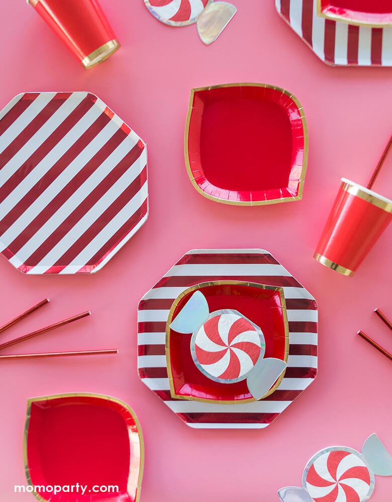 Pink & Red holiday party table set up with Jollity & Co Red 6" Christmas Peppermint Small Napkins, Posh Ruby Kiss Dessert Plates, Red stripe dinner plates, Posh Ruby Kiss Cups with red party paper straw on a pink background, for a sweet holiday celebration