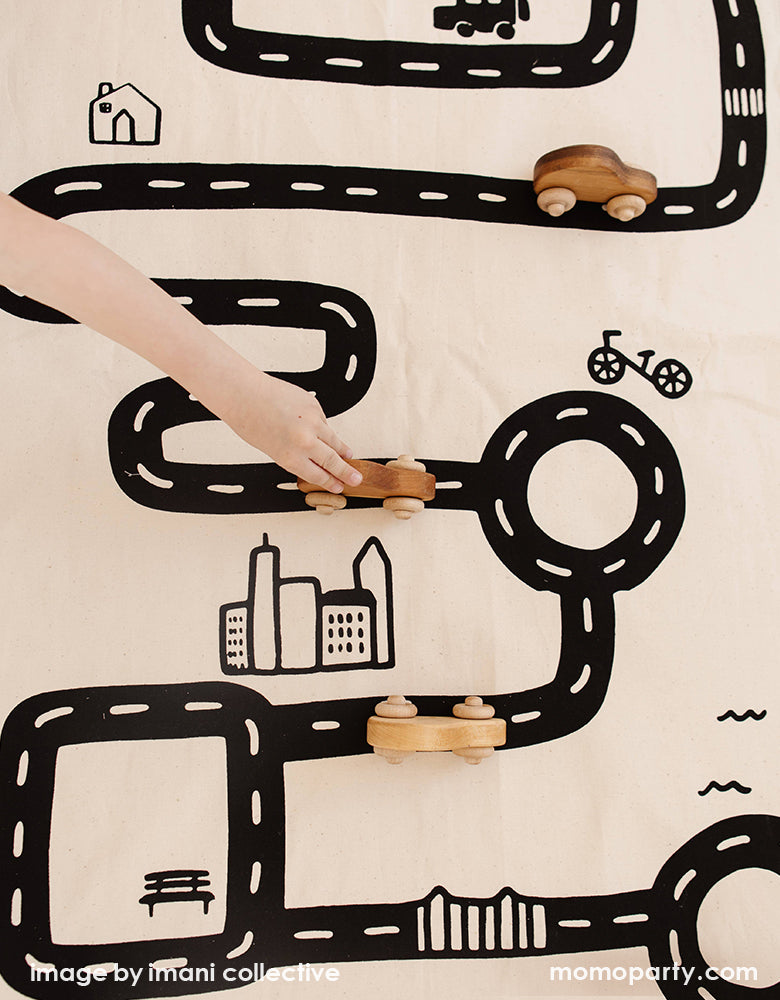 A top view look of A Boy's arm play with his wooden car toys on Imani Collective interactive play floor mat - Road Floor Mat. This floor mat is natural canvas hand with black screen printed road bike, houses modern illustration, Sewn and screen printed by hand on natural canvas by Kenyan artisans. Sold by Momo party store provided modern party supplies, boutique party supplies