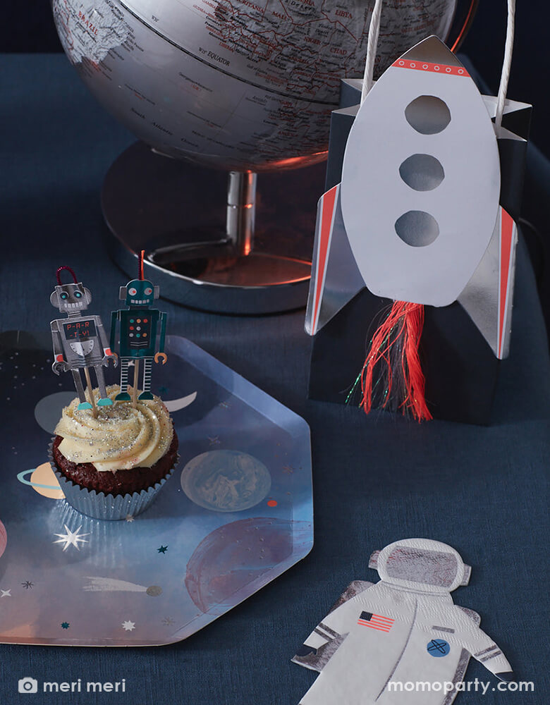 Close up of space themed party table with\ cupcakes decorated with space cupcake toppers on a Space Dinner Plate by Meri Meri, Astronaut Napkins, Rocket Party Bag, and a silver globe. These modern unique designed party supplies are perfect for a space themed birthday party, blast off birthday party, two the moon birthday party.