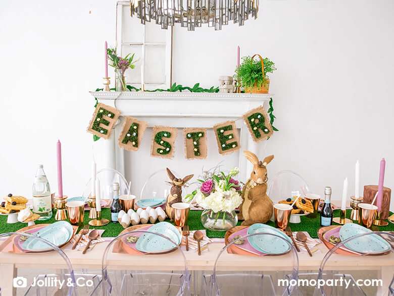 Easter party table set up with Rockin' Robin Dinner Plates layered with pink posh plates, fresh flowers and bunny figures, eggs, drinks, pink candle on a grass feel table runner, a nurture cloth with artificial grass letter in "EASTER" text with mini eggs banner on the back