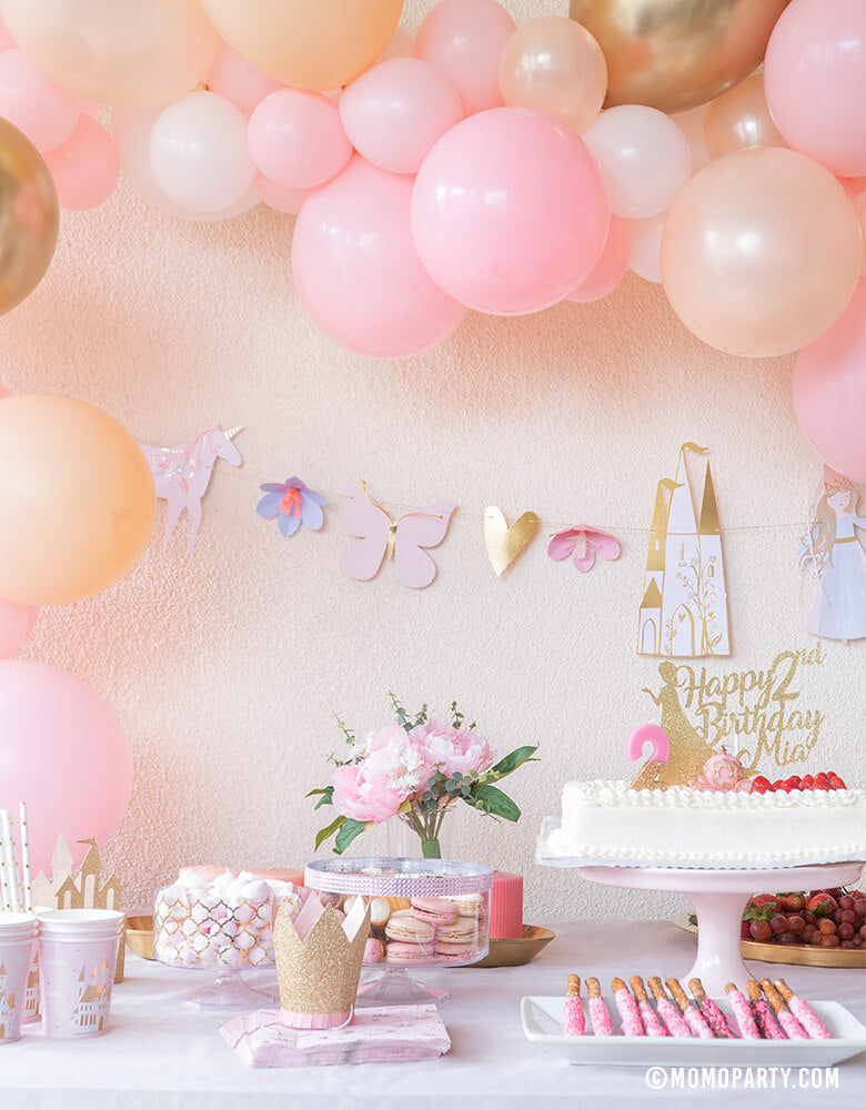 Pink and gold Modern Royal Princess Party dessert table close up look, decorated with Pink Gold Peach color mixed balloon garland, Meri Meri Magical Princess Garland , cake with customized gold glitter princess topper, flowers, princess castle paper cups, crown hats, soft pink cotton candy in a pink container, fruits for a girls pink gold princess themed birthday party celebration