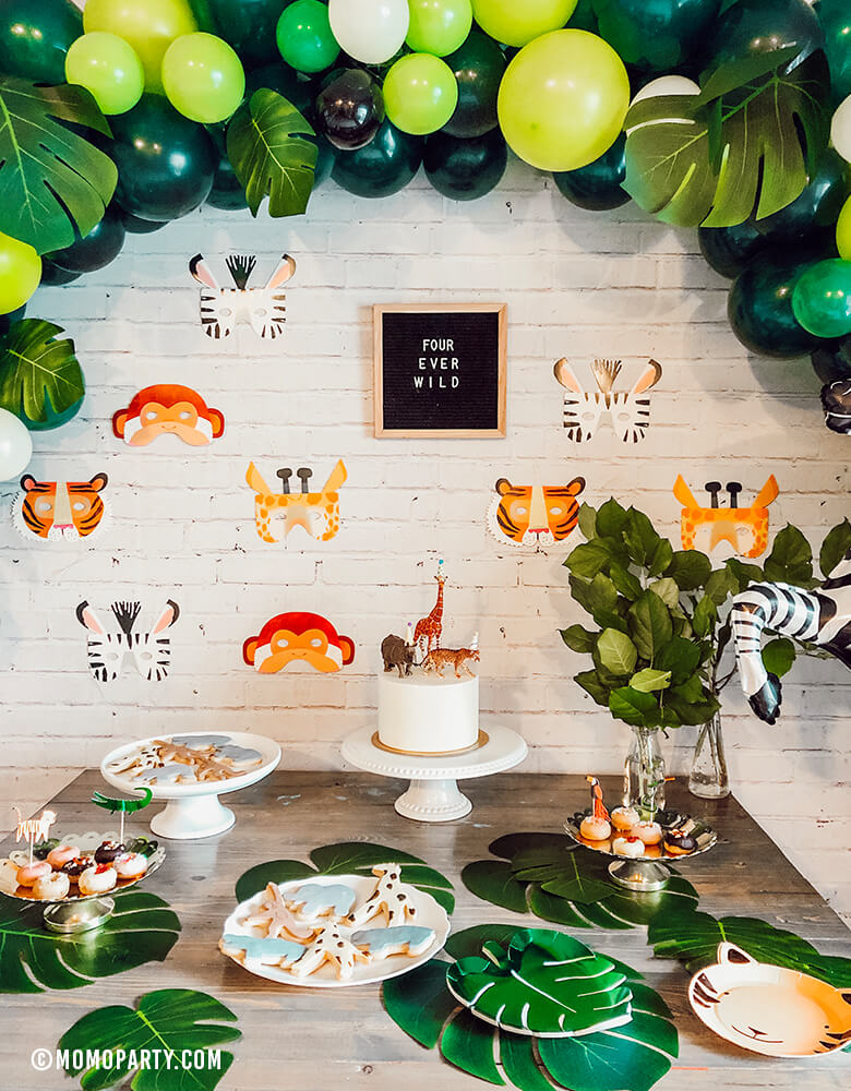 Safari Jungle Birthday Party desert table with safari themed balloon garland and Talking Tables, Party Animals Paper Masks of Zebra, Giraffe, Tiger and Monkey and letter board of "Four ever wild" sign as backdrop wall decoration.