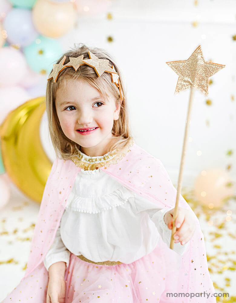 Little girl wearing a pink star sparkle cape Costume and gold Shining Star Headband, holding a Shining Star Wand just like a magical princess in a party celebration 