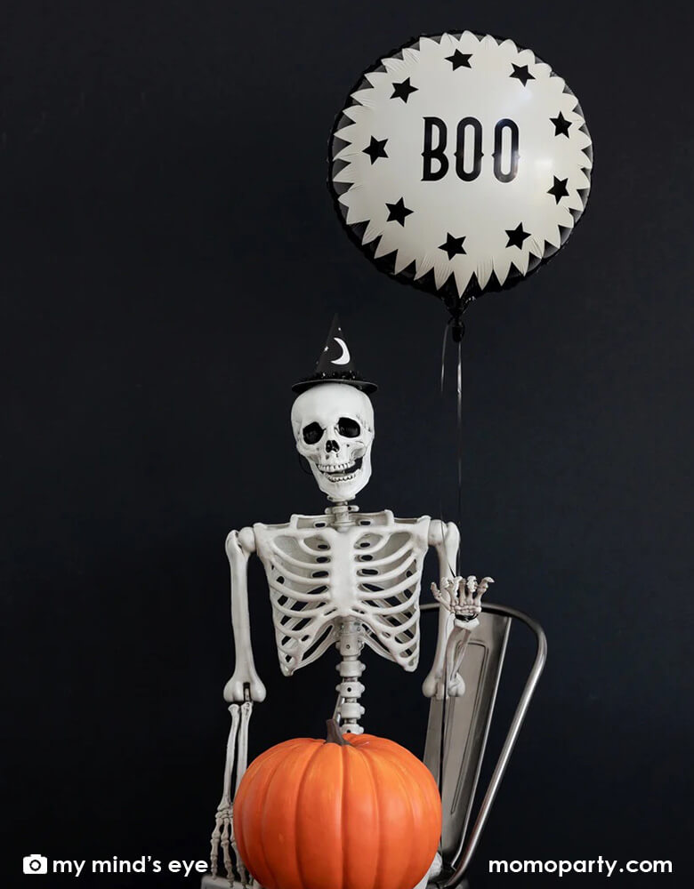 A posable Halloween skeleton holding a pumpkin in his lap and on his back there is My Mind's Eye's Vintage Halloween Boo Foil balloon floating, creating a spooky vibe for a Halloween decoration look