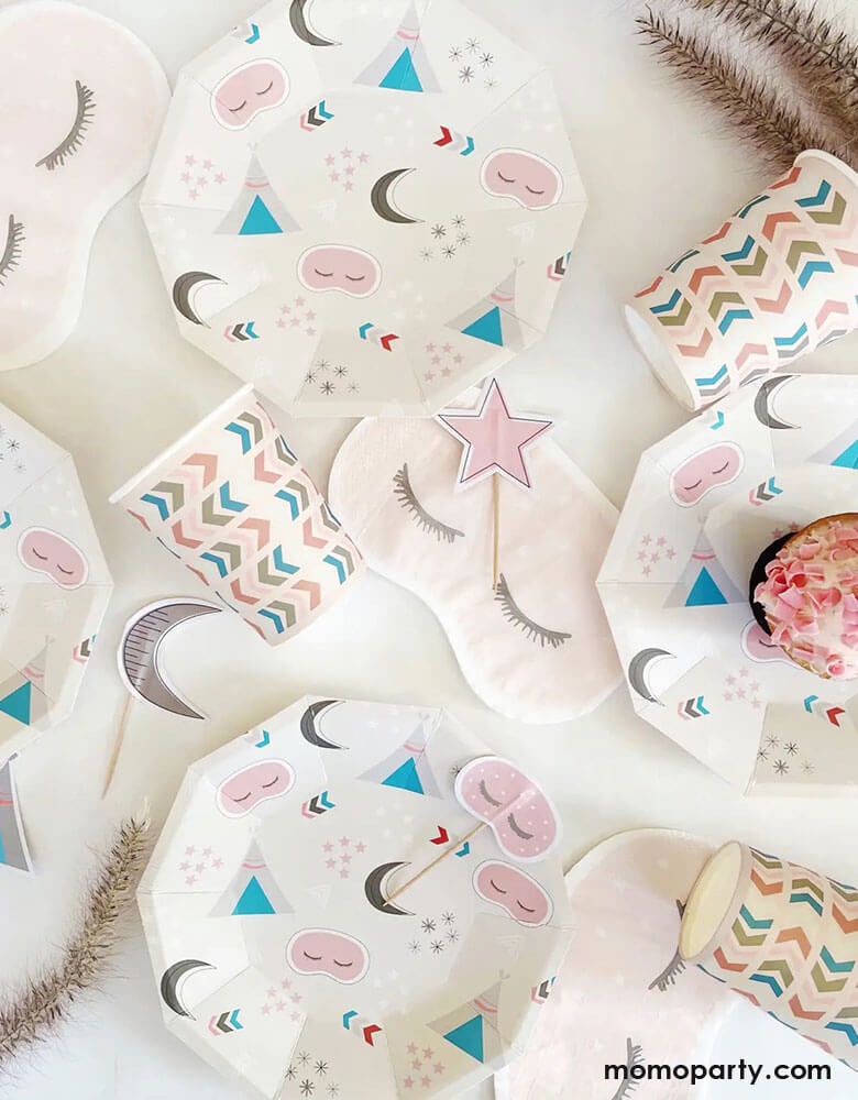 A tablescape of girls sleepover party featuring Momo Party's sleepover napkins in the shape of sleeping masks by Pooka Party, along with coordinated plates featuring sleepover themed illustrations like sleeping tents, sleeping masks, moons and stars, plus light pink party cups with chevron design, and cupcake topers with moon design, perfect for girl's slumber party.