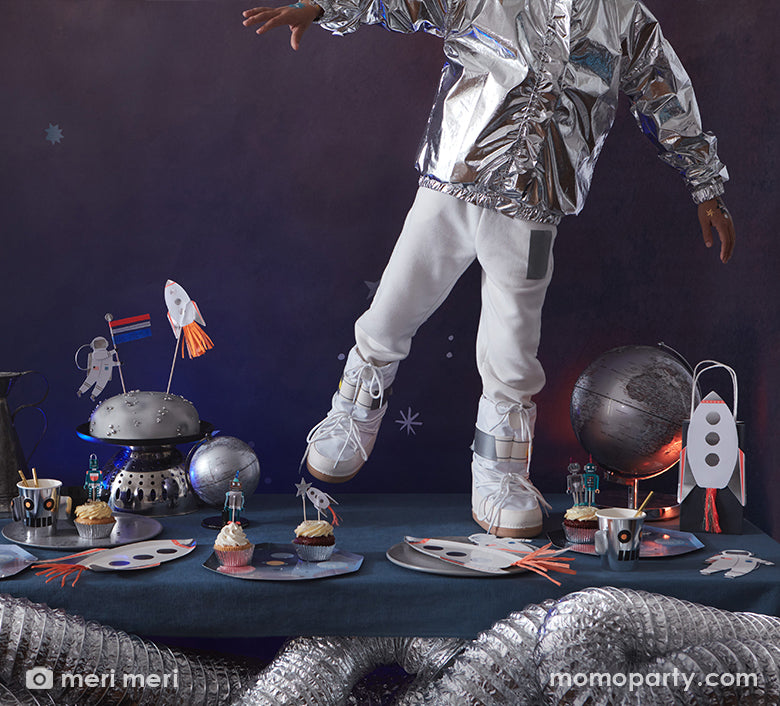 A kid wearing a space suit standing on his space themed birthday party table, filled with Rocket Plates by Meri Meri, cupcakes decorated with space cupcake toppers on a Space Dinner Plate, Robot Cup with gold straws, silver moon cake set decorated with cake toppers, Rocket Party Bags, a silver globe. These modern unique designed party supplies are perfect for a space themed birthday party, blast off birthday party, two the moon birthday party.