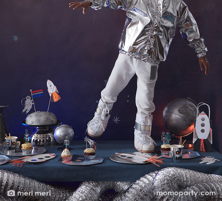 A kid dressed like an astronaut in his space suit standing on a party table filled with Meri Meri's space themed party supplies including space dinner plates with planets design, rocket ship shaped plates, astronaut shaped foil balloon and a moon inspired birthday cake topped with space themed toppers 