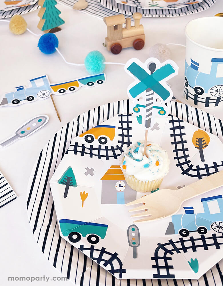 Boy's Train themed birthday party table set up with Pooka Party Steam Train Small Plates layered with Black & White Fine Stripes Large Plates. A cupcake decorated with a stop sign topper from Steam Train Cupcake Toppers kit. There are Steam Train Cups, wooden train toys, pom pom garland around the table. These steam train partware collection featuring blue, green and orange colors with train related design elements like train, train truck, stop sign. It will make your train themed party the most stylish.