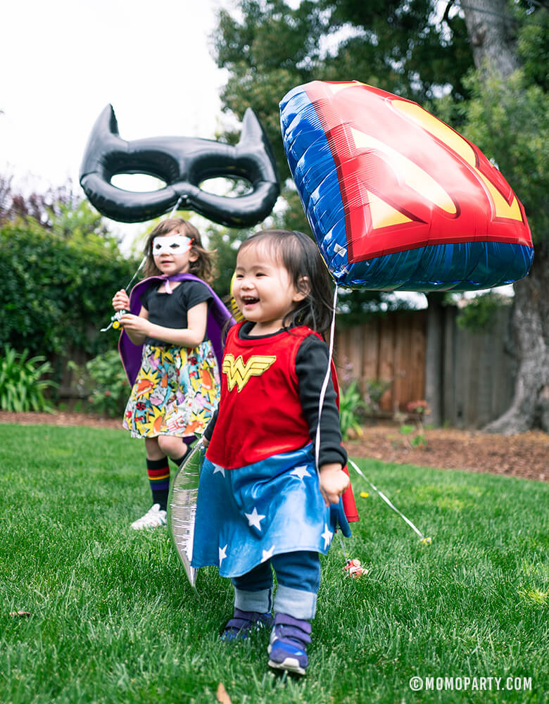 little girl wear wonder woman costume holding a Superman Emblem Foil Balloon with another girl holding giant black bat mask shaped foil balloon in a kid superhero birthday party