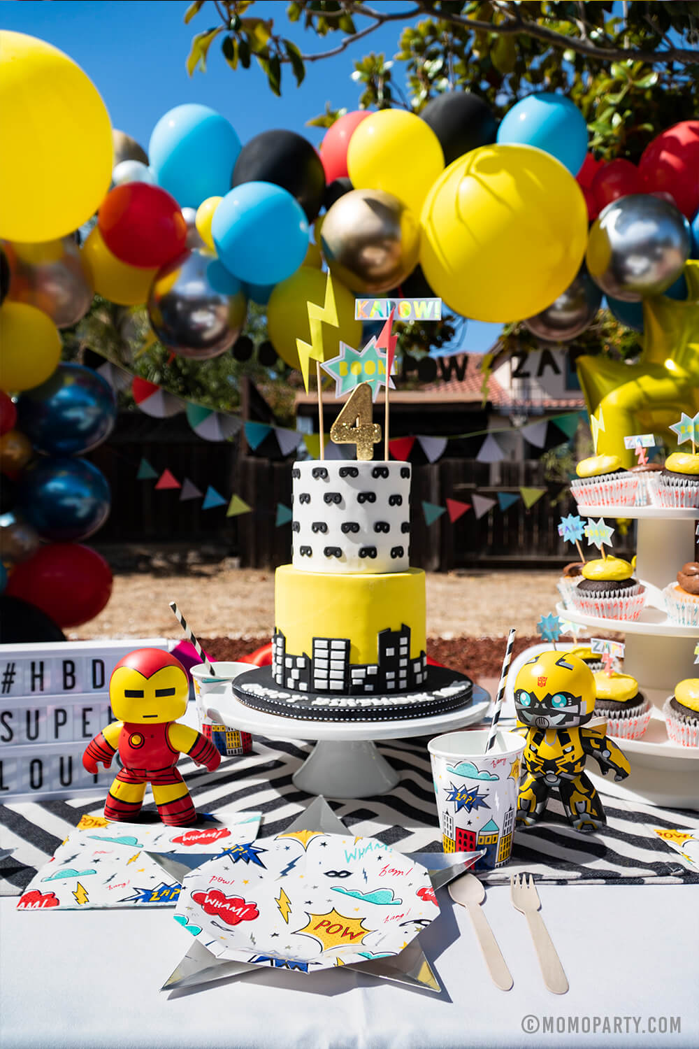 Kids modern superhero birthday party table set up with Day Dream Society Superhero Party Small Paper Plate, Star Silver Foil Plate, Superhero Napkins, and wooden utensil, Modern Superhero cake with number 4 gold glitter candle, superhero sign cake topper, balloon garland as decoration on the back