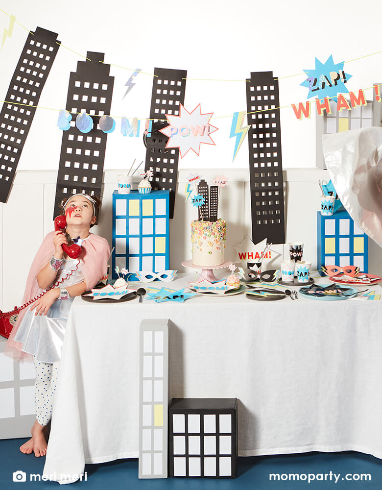 Kids superhero party with a girl costume, lighting face painting, holding a red telephone standing next to a superhero themed party table with superhero comic word shaped plates, superhero cups, neon Lightning Bolt Napkins, a colorful chorios cake decorated with superhero cake toppers, cupcake with superhero themed toppers, superhero paper masks on the table, and a large superhero garland hanging on the wall. Such a fun, bold and modern superhero themed birthday for any superhero lovers