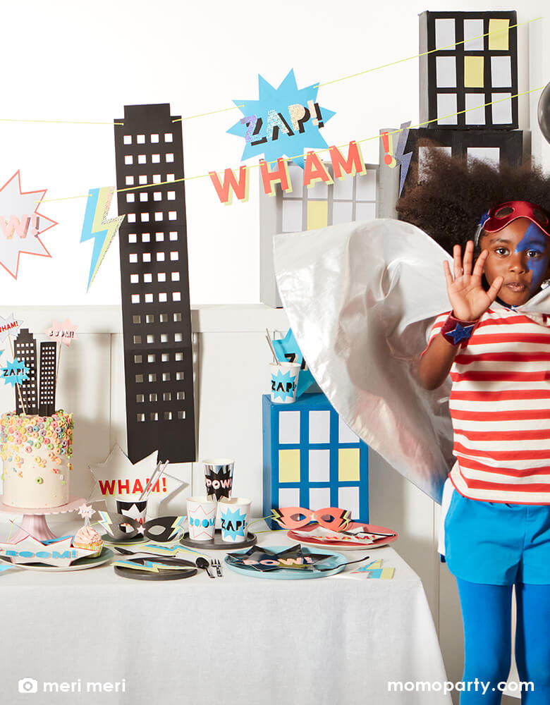 Kids superhero party with a girl wearing a cape and mask, dancing next to a superhero themed party table with superhero comic word shaped plates, superhero cups, neon Lightning Bolt Napkins, a colorful chorios cake decorated with superhero cake toppers, and a large superhero garland hanging on the wall. Such a fun, bold and modern superhero themed birthday for any superhero lovers