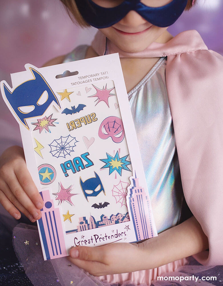 Girl wearing a blue silk mask and pink cape, holing a Great Pretenders - Superhero Star Temporary Tattoos. This set of temporary tattoos includes bats, spiders, and superhero emblems made with a glitter finish to reflect beautifully on the skin! It makes great party favor for your super girl's guests! fun activity for superhero themed party, girl power party or any dress up party