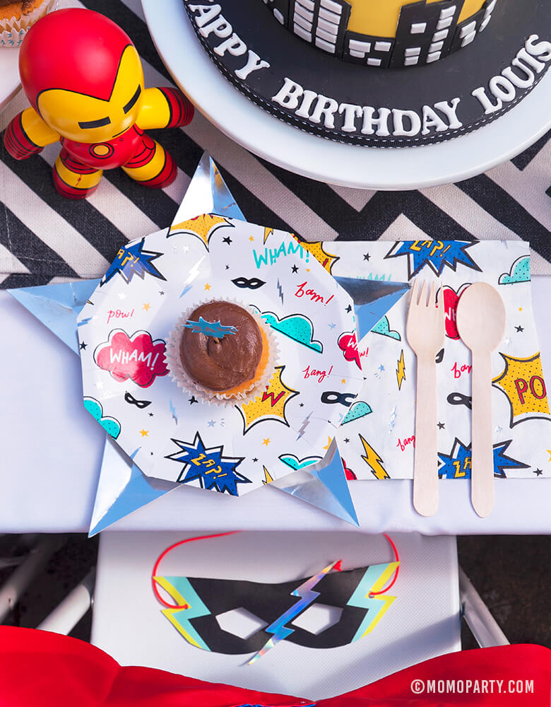Kids modern superhero birthday party table with Day Dream Society Superhero Party Small Paper Plate, Star Silver Foil Plate, Superhero Napkins, and wooden utensil, a Superhero Mask on the chair