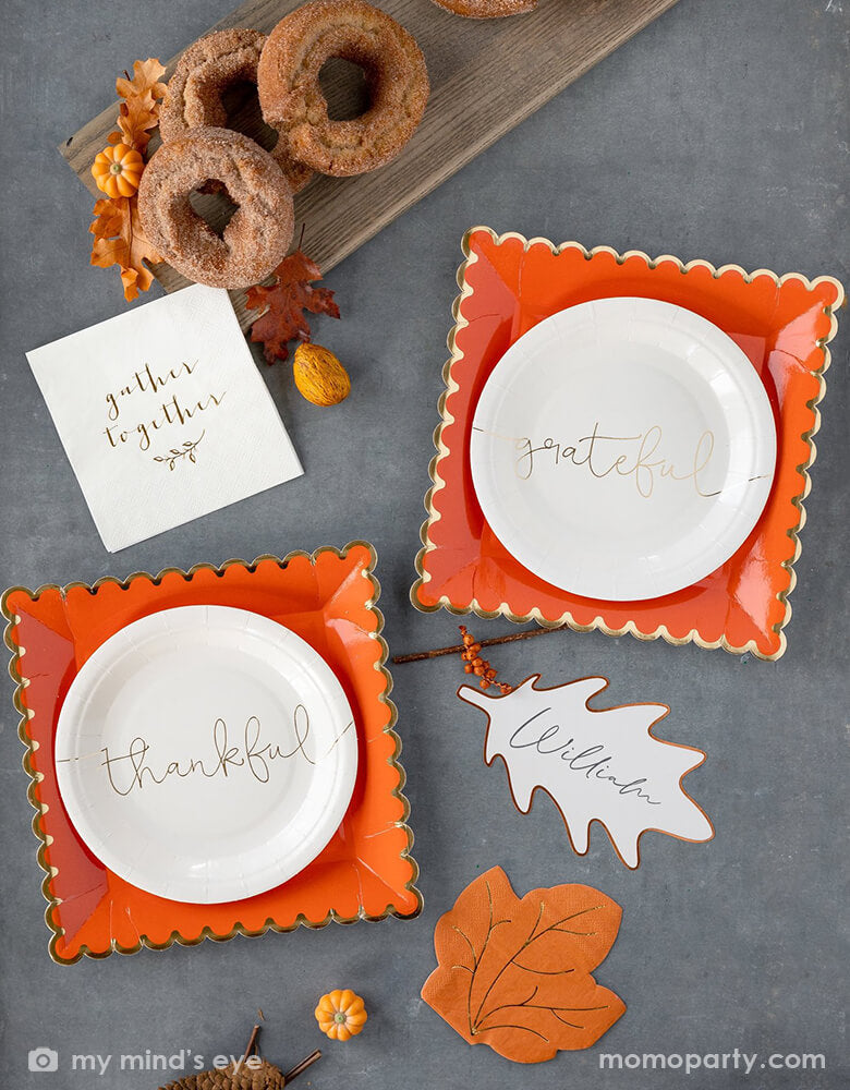 Table top view of My mind's Eye 7 inches Thankful Grateful Plates with two "thankful" and " Grateful" gold foil script printed cream paper plates, layered with Orange with golden foil scalloped large plates, there are cinnamon donuts with mini pumpkins decoration on a wooden cheese board, a maple leave napkin, and gather together napkin, dry Maple leaves, nuts all around on top of the grey quartz countertop. These modern party supplies are Perfect for your Thanksgiving celebration dessert or appetizers.