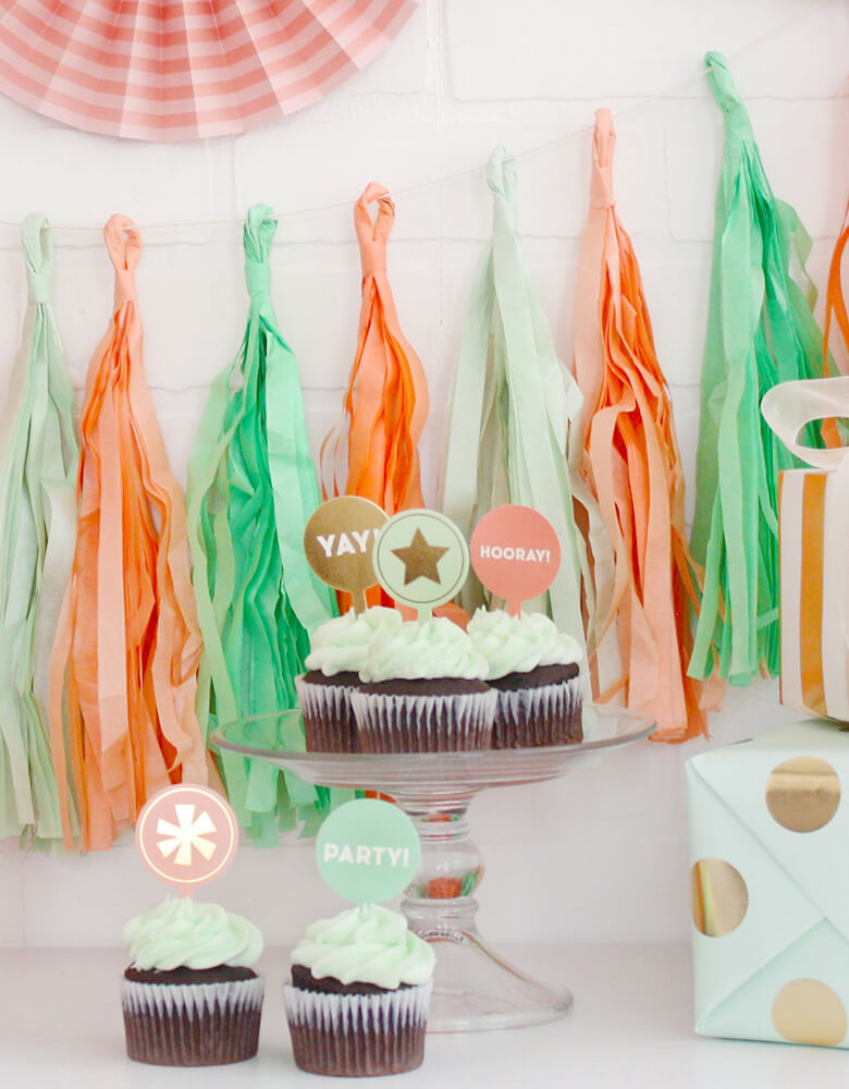 A modern Birthday party with Trend Tassel Banner in peach, mint and coral colors hung on the wall and mint cupcakes with "yay" "Hooray" "Party" cupcake toppers on the table with birthday gifts wrapped in mint dotted paper