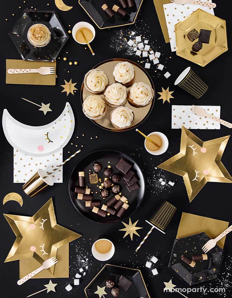 A festive new year eve table with black table cover filled with black and gold party goods including die-cut little star plates in gold, little moon plates in white and black cups and plates with gold accent
