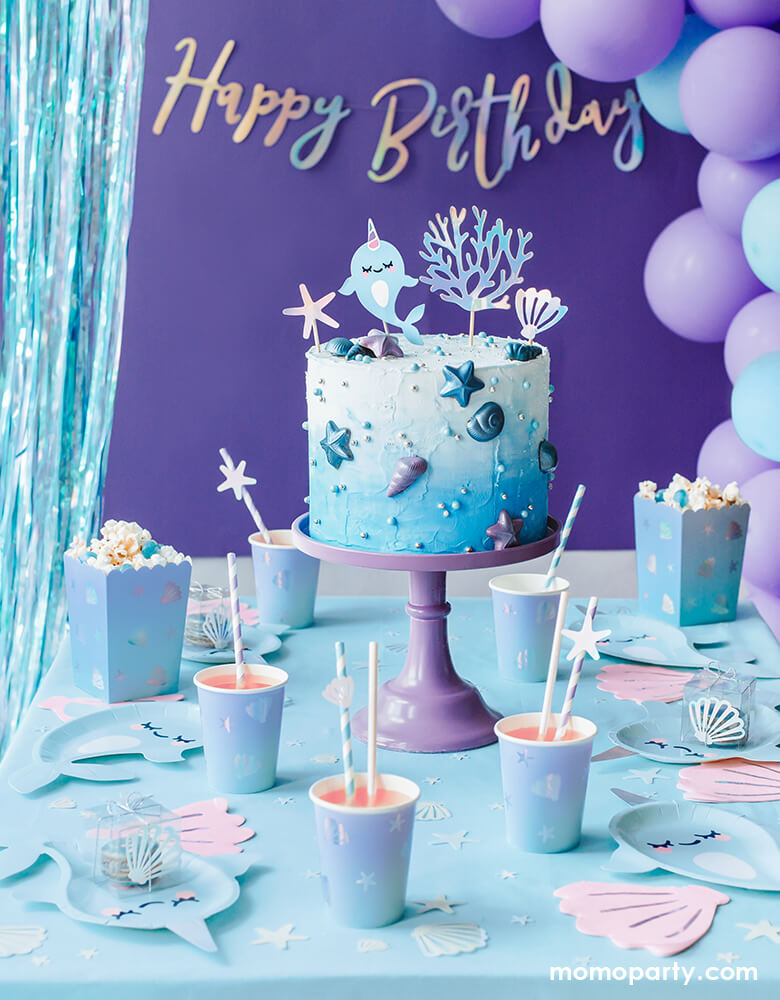 Under The Sea Narwhal Party Table look, with a iridescent happy birthday garland, blue Party curtain, and purple light blue mixed balloon garland as decoration, there is a ombre blue buttercream cake decorated with Narwhal cake topper in the middle of the table, Under The Sea Ombre Cups with paper party straws, Narwhal paper plates, Pink Shell Napkins, Under The Sea Popcorn Treat Boxes with popcorns, iridescent confettis on the table. Super dreamy party set up for a under the sea birthday party