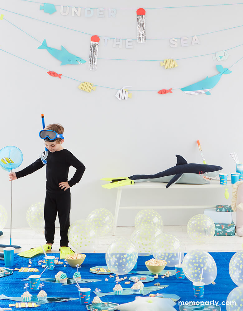 Boy wearing his Divers Snorkeling Set, holding a net with a fish napkin inside, with Meri Meri Under The Sea Garland on the wall, big shark toys wearing Divers Snorkeling Set as well, with lots of balloons on the floor with a big rug, sea cups, plates, shark platters, sweets, so fun for a sea themed party, shark themed birthday party, under the sea birthday party, boy themed birthday party, or any celebration for sea lover