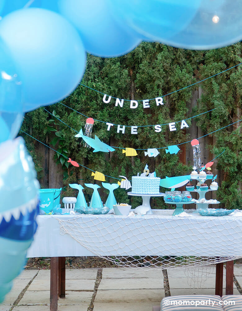 under the sea birthday party with Meri Meri Under The Sea Garland with shark, fish, whale and jellyfish hanging on the tree. Cake with number 6 candle, shark hats, cupcakes on the blue cake stand on the table with net decoration for a boy's 6 years sea friends themed  birthday party 