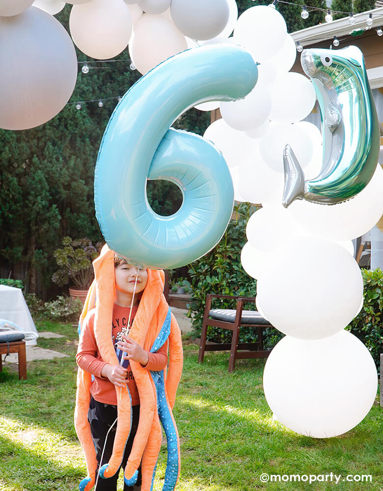 Outdoor Under the sea themed birthday party of boy wearing a octopus costume and holding a Large Number 6 Pastel Blue Foil Mylar Balloon with Hammerhead Shark Foil Balloon and balloon garland on his back, happy celebrating his modern shark themed birthday party