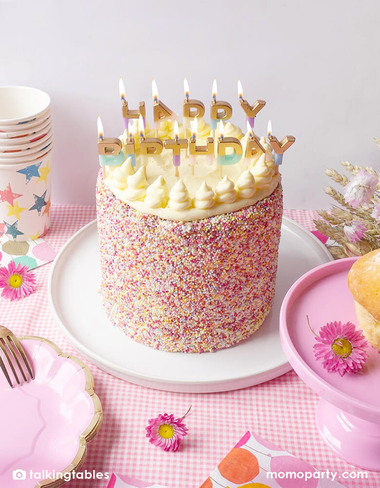 A birthday party table with a red-pink tie sprinkle cake on a round plate, decorated with decorated in pastel colors and half-dipped-glitter We Heart Birthdays Happy Birthday Candles by Talking Tables. Surrounded by cupcakes on a pink cake stand, flowers and colorful stars cups, they are all on a pink checkered tablecloth, ready for birthday party