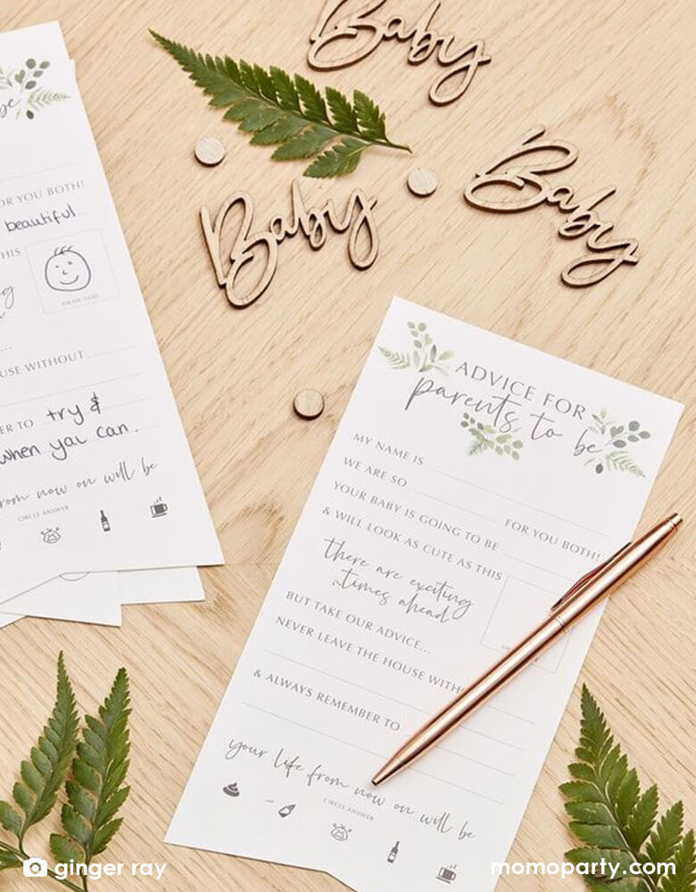 Modern baby shower table close up with Ginger Ray - Wooden Baby Confetti next to some "Advise_for_parents_cards", green leaves on a wooden table. This Wooden Baby Confetti Contains 18 Wooden Confetti Pieces of "baby" script shape and small circle pieces. Scatter your table with this wooden baby shower confetti to compliment a natural baby shower table decorations.
