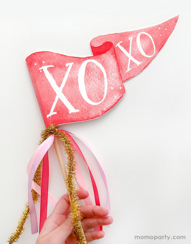 hand holding Cami Monet - XOXO Party Pennant. This Handmade pennant made in United States of America, in Size: 10 x 5 inches. This is made of 120 lb. luxe watercolor texture paper with handwriting "XOXO" text in watercolor illustration for extra whimsy. With pink and red and gold mixed Ribbon and sparkle garland. This adorable party pennant is a perfect for a sweet Valentine's Day celebration!