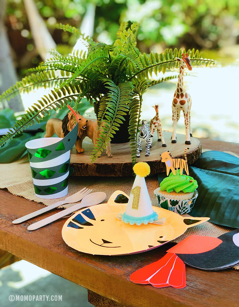 Safari Jungle Outdoor Party Table Set Up with Tiger Paper Plates, Toucan Napkins, Snake paper cups, animal figure toys, get wild cupcake kit, 1 year birthday hat on a wooden picnic table.