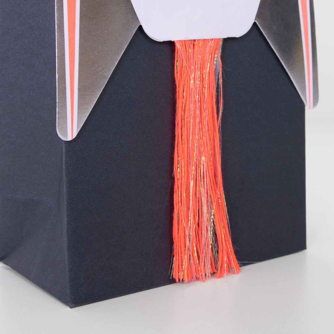 A detail close up with Rocket Party Bags by Meri Meri. There are beautifully embellished with shiny silver foil and a shimmering tassel on a dark navy paper pag. with Thick neon coral and fine iridescent thread embellishment. Your guests will be delighted to zoom off home with these awesome rocket party bags.