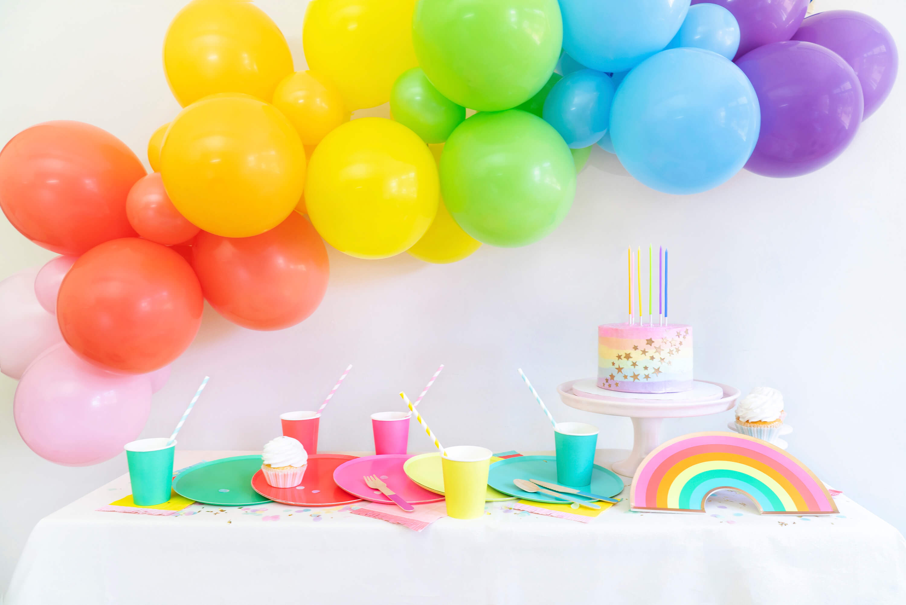 Momo party Modern rainbow party table look with colorful rainbow large paper plates, cups, pastel rainbow cake with rainbow candles, rainbow balloon garland with pink, coral, orange, yellow, lime, blue and purple latex balloon