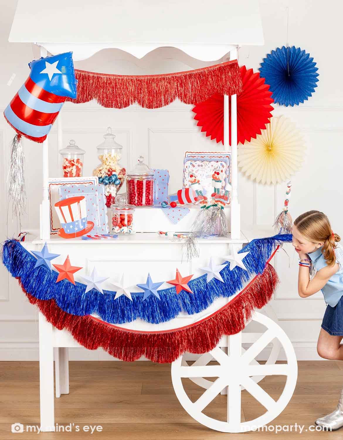 A Fourth of July themed decorated dessert cart with various American themed decors and tablewares in red, white, and blue from Momo Party, including paper fans, fringe bunting garlands, star party banners, a firecracker shaped foil balloon, plates, napkins and cups, along with a girl in blue shirt and skirt standing next to it. A perfect inspiration for Fourth of July, Memorial Day, or summer celebrations with kids.