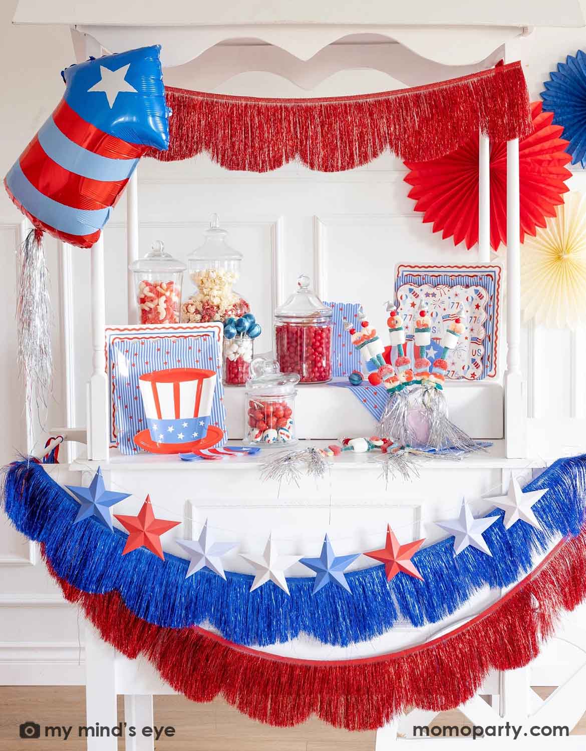A close up of a Fourth of July themed decorated dessert cart with various American themed decors and tablewares in red, white, and blue from Momo Party, including paper fans, fringe bunting garlands, star party banners, a firecracker shaped foil balloon, plates, napkins and cups. A perfect inspiration for Fourth of July, Memorial Day, or summer celebrations with kids.