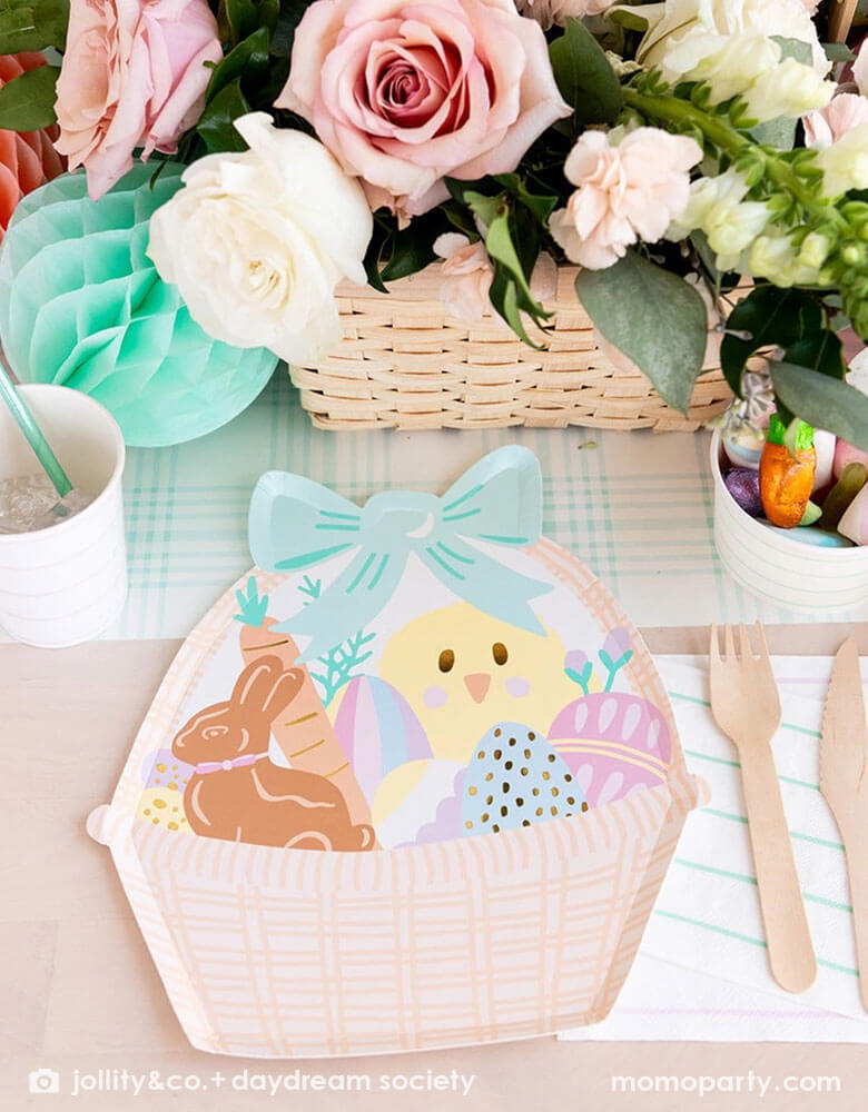 An adorable kid's friendly Easter table setting features Momo Party's Easter Fun basket shaped plates, mint striped plates, party cups by Daydream Society. In the middle there's light blue gingham table runner, Easter Egg tissue honeycomb in mint and peach and a beautiful Easter flower arrangement in a basket. On the table there's also a small treat cup filled with Easter candies and chocolates, making this an adorable Easter tablescape.