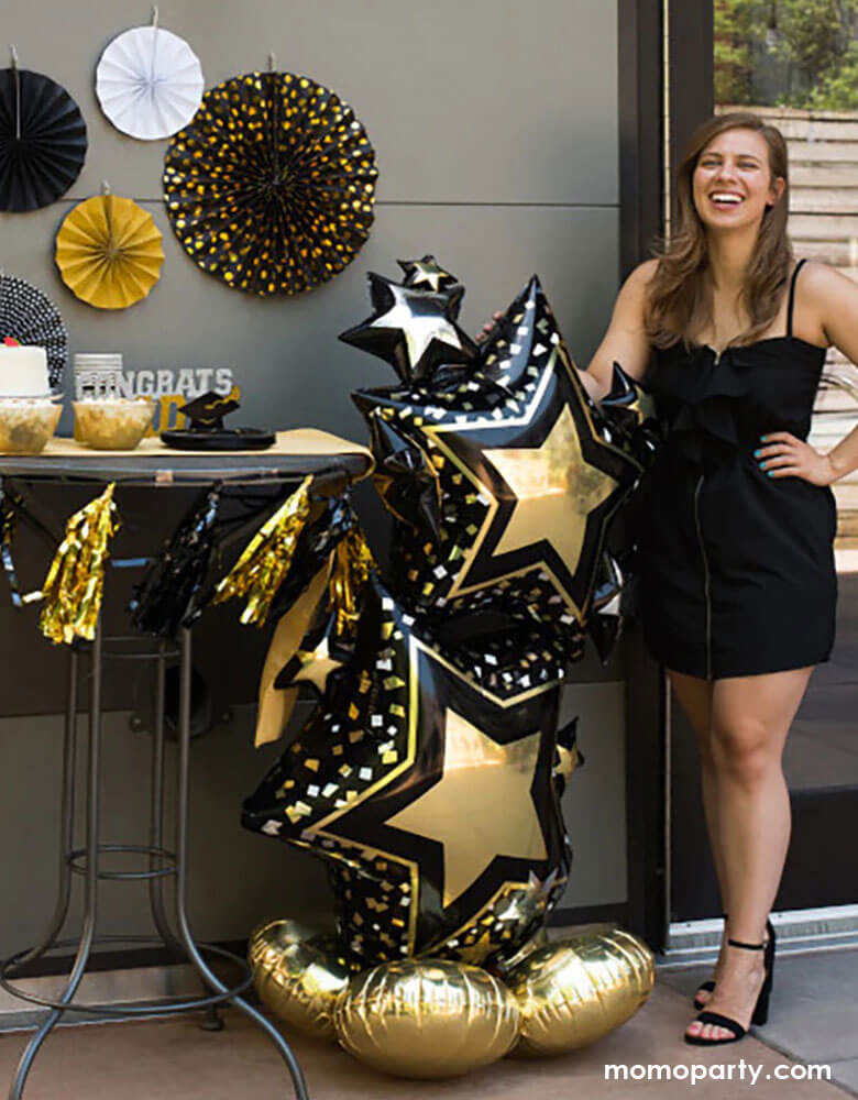 A collage or high school graduate standing next to Momo Party's 59" Airloonz Black and Gold Star Cluster Foil Balloon by Anagram Balloons next to a dessert table decorated with graduation themed decorations including black and gold party fans, tassels, "Congrats" candles to celebrate her graduation day.