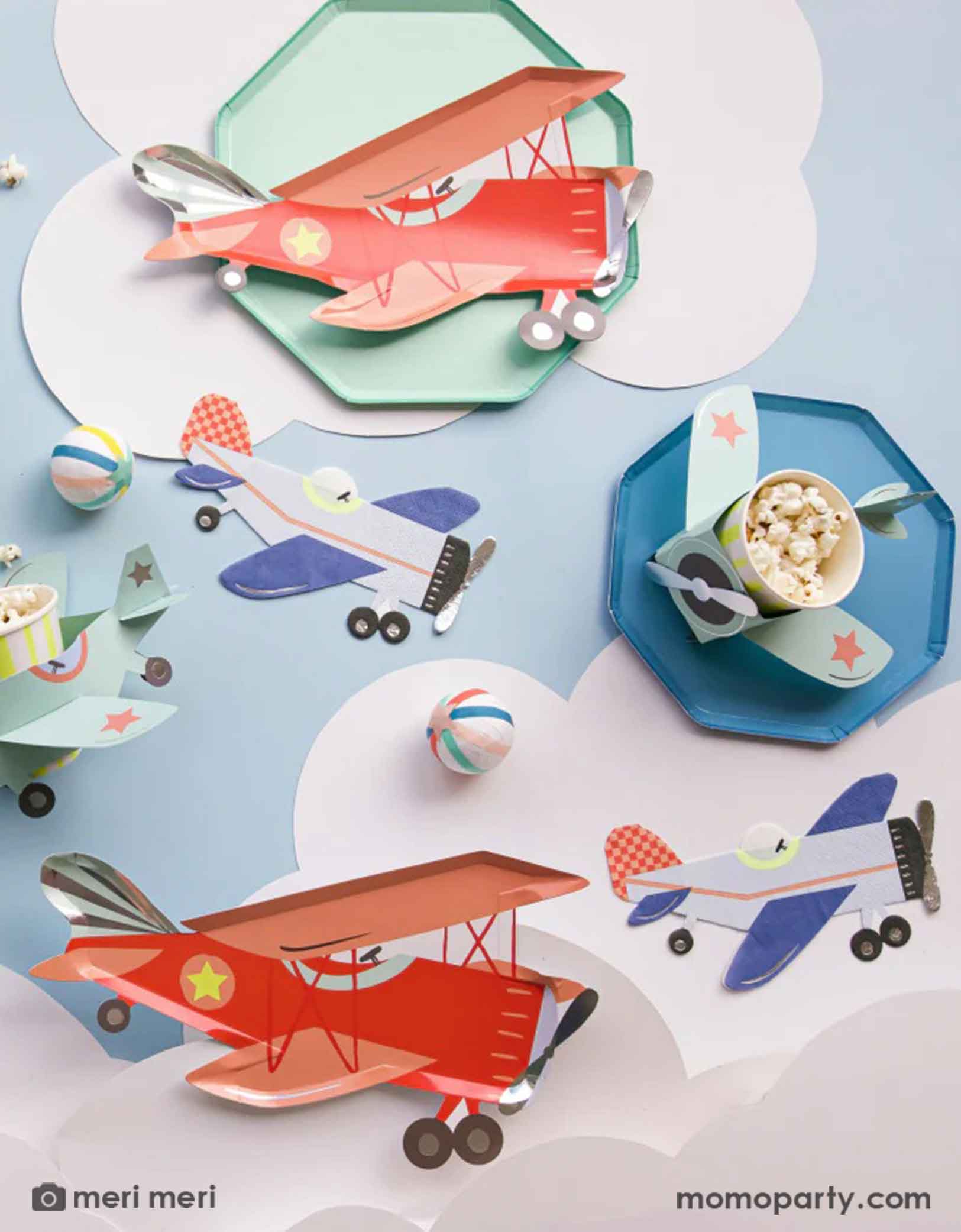 A table of fun and adorable airplane themed party tableware from Momo Party by Meri Meri, including red retro vintage plane shaped paper plates, blue vintage plan shaped napkins and airplane 3D paper party cups, along with cloud paper cutouts, this look is a great inspiration for kid's airplane themed party decorations.