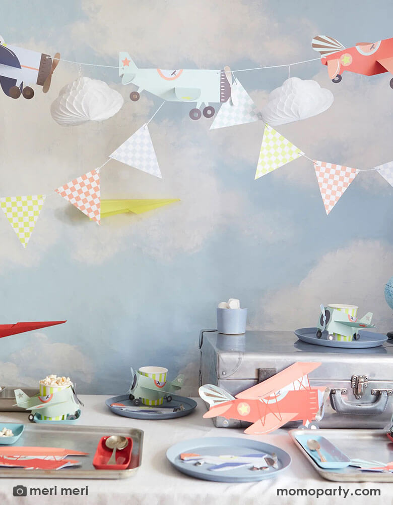 A airplane themed birthday party featuring Momo Party's plane themed decorations and party supplies by Meri Meri including red vintage airplane shaped plates, blue airplane shaped napkins and paper cups filled with drinks and treats. Above the table is a 3D honeycomb paper crafted plane garland against a blue sky with clouds wallpaper which sets a perfect scene for this airplane / aviator themed birthday party.