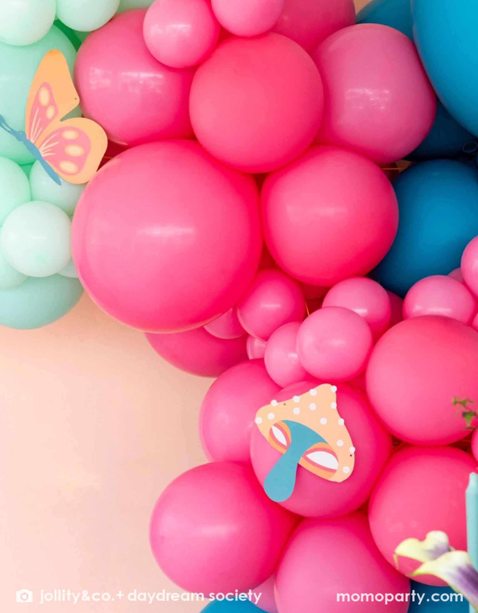 A colorful spring inspired balloon arch with pink, blue, and green balloons adorned with butterfly and mushroom paper pennants from Momo Party's Alice Dreamland Whimsy Garland Set on a light background.