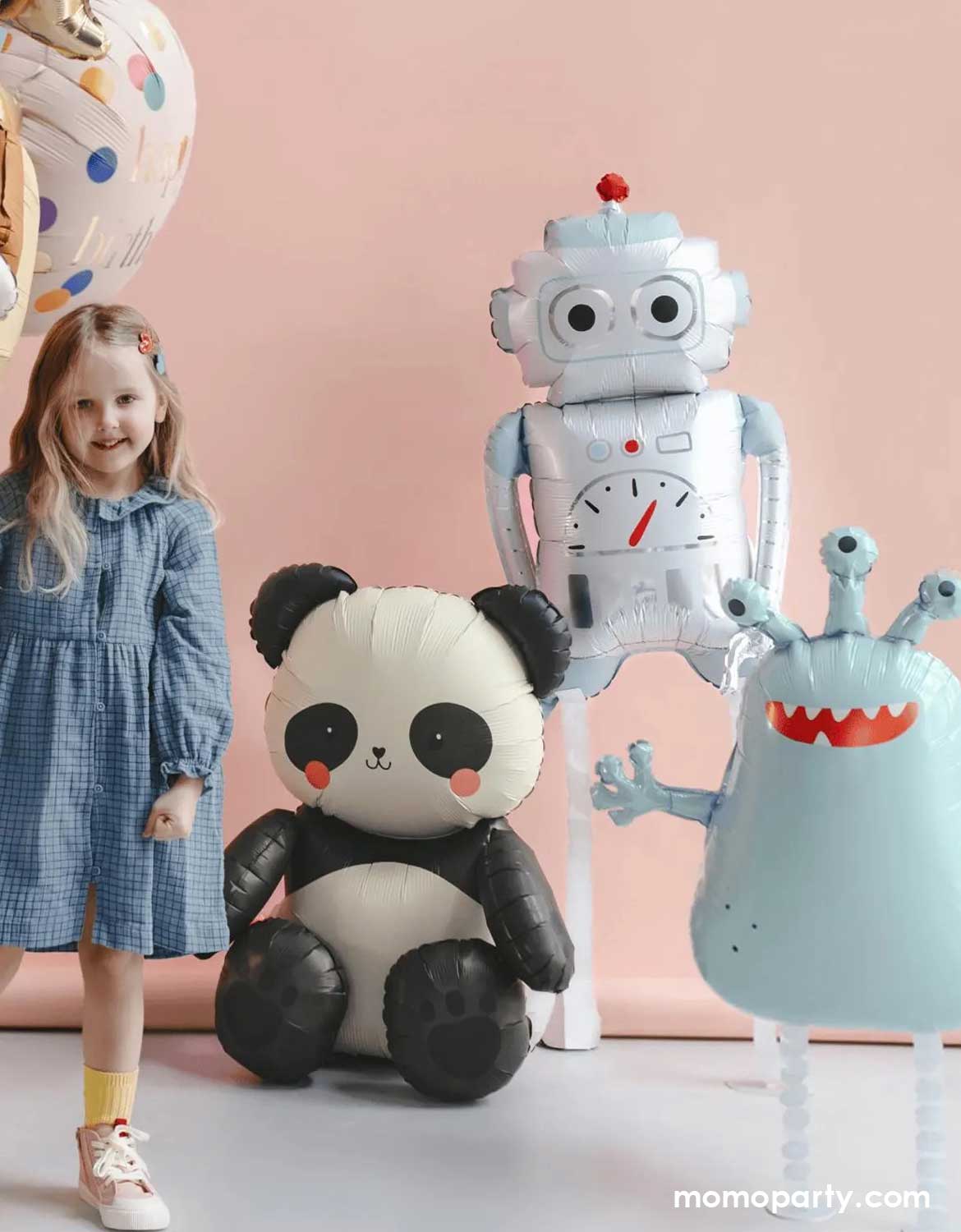 A girl with various large foil balloons around her from Momo Party including a sitting panda shaped balloon, a standing silver robot balloon and a mint alien shaped monster foil balloon against a light pink wall.