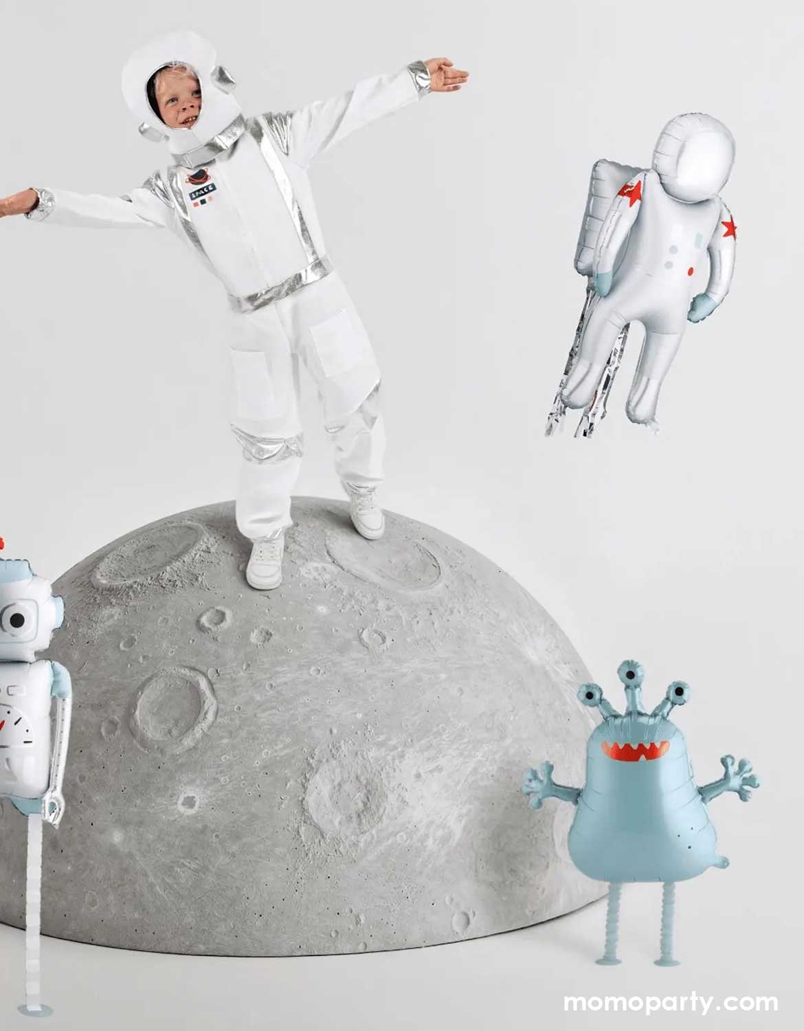 A child in astronaut costume standing on a moon surface with space-themed foil balloons from Momo Party including an astronaut shaped balloon, a robot balloon and a mint alien shaped balloon.
