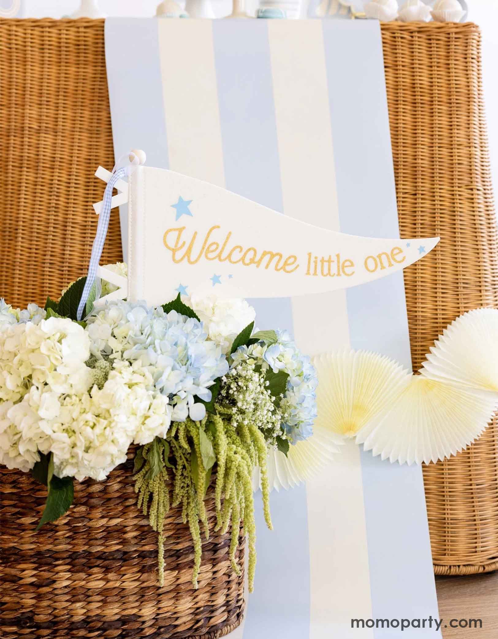 A modern and clean boy baby shower set up featuring straw-strung table covered with Momo Party's baby blue and white striped table runner with flowers in a basket and 'Welcome little one' sign at the bottom.