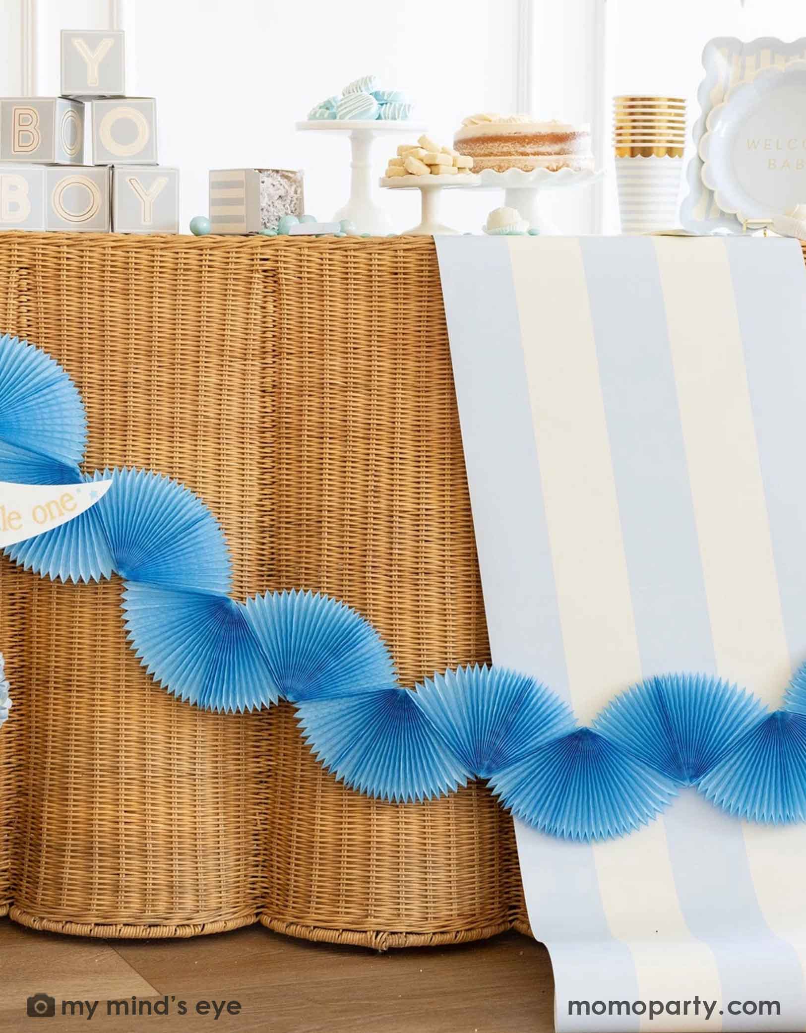 A modern and clean boy baby shower set up featuring straw-strung table covered with Momo Party's baby blue and white striped table runner. On the table there are various baby shower tablewares from Momo Party including party cups, treat boxes, plates and more. There's also a blue bunting banner hung across the table.