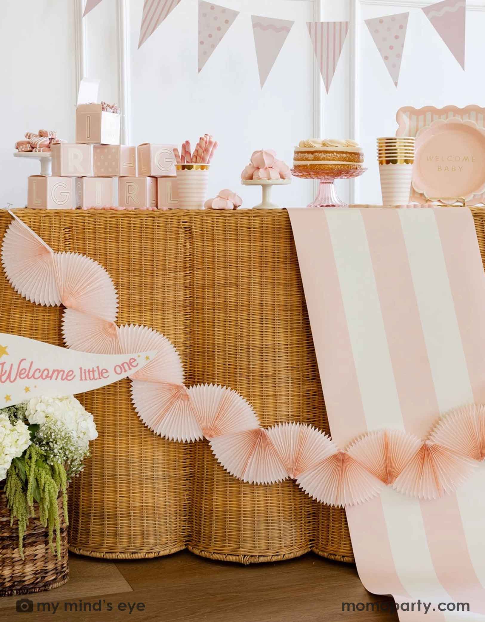 A modern and clean gril baby shower set up featuring straw-strung table covered with Momo Party's baby pink and white striped table runner. On the table there are various baby girl shower tablewares from Momo Party including party cups, treat boxes, plates and more. There's also a pastel pink bunting banner hung across the table and a "Welcome Little One" party pennant in the flower basket at the bottom.