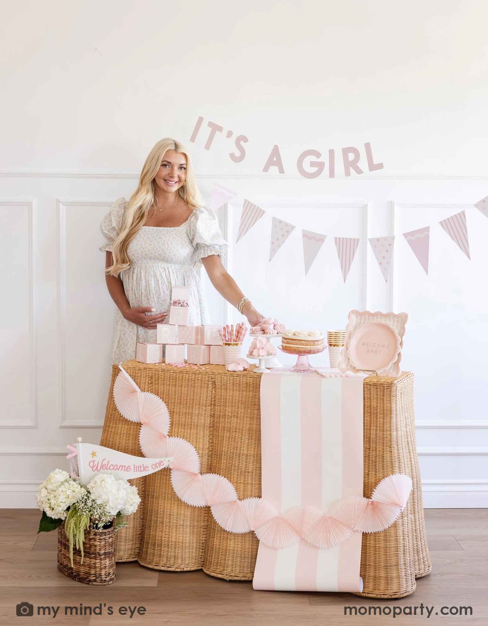 A pregnant woman standing next to a baby shower table with 'It's a Girl' banner and baby pink shower decorations and party supplies from Momo Party including tablewares, party banners, table runner and bunting garlands.