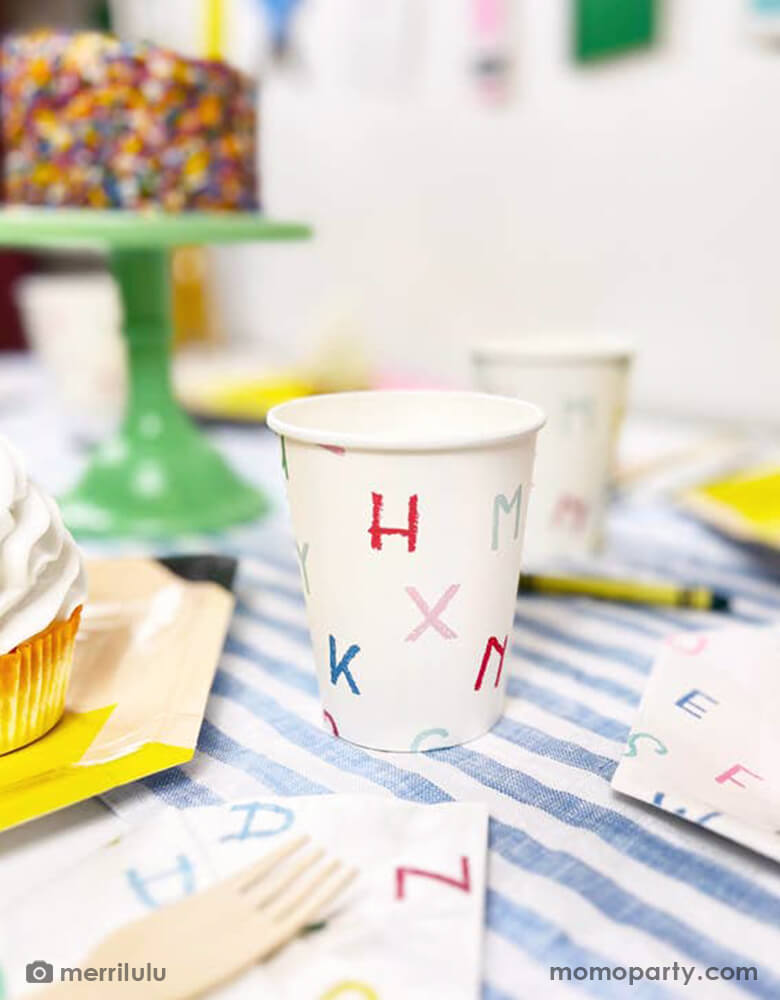 A close up shot of a back to school party table featuring Momo Party's alphabet letter patterned party cups by Merrilulu. In the back you can see Next to the tableware there's a cake topped with a pencil shaped cake topper with "Hello 2nd Grade". In the back there's a back to school party banner featuring colorful school themed pennants including crayon, glue bottle, notebook and more. All this makes a great inspo for a back to school party or first day of school celebration.