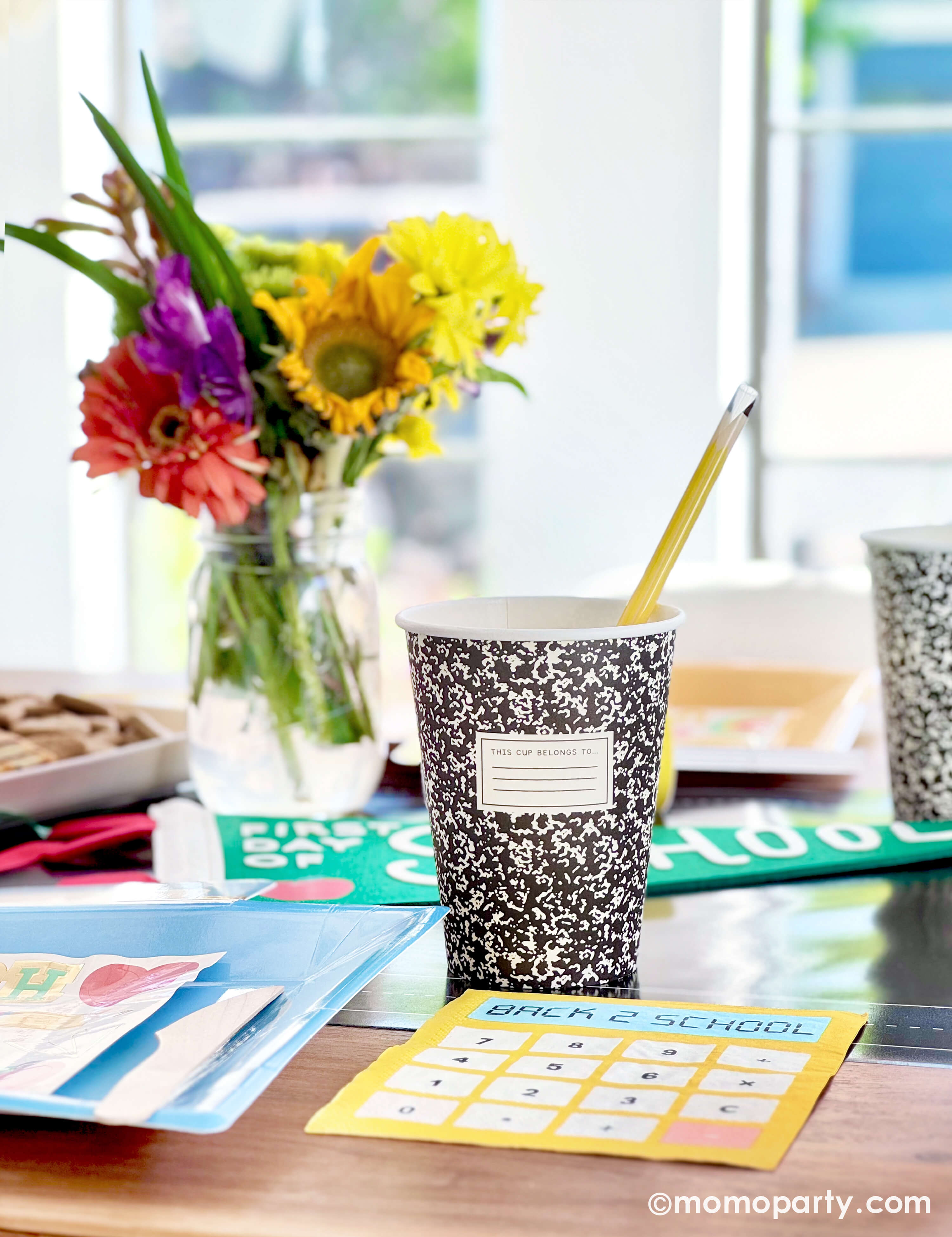A kids' back to school party table setting featuring various school themed party decors and tableware from Momo Party including notebook party cups with pencil reusable straws, calculator shaped dinner napkins, lunchbox shaped plates and apple first day of school party pennant, with a summer flower arrangement and a kid's snack board, along with "School Day" party garland, this simple yet festive set up is perfect to celebrate your child's first day of school!