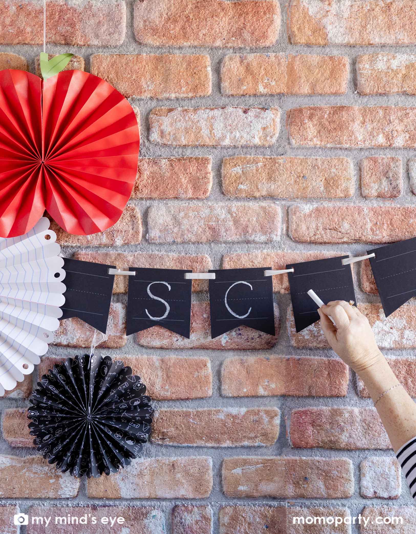 A brick wall decorated with Momo Party's back to school decorations including the Chalkboard Writable Paper Banner and the back to school party fans featuring an apple shaped fan and notebook patterned party fans. A hand is writing SC on the write-on party banner with a chalk. This banner lets you customize messages with chalk or chalk markers. Easy to write on and erase, it adds a fun, interactive touch to any celebration or learning space.