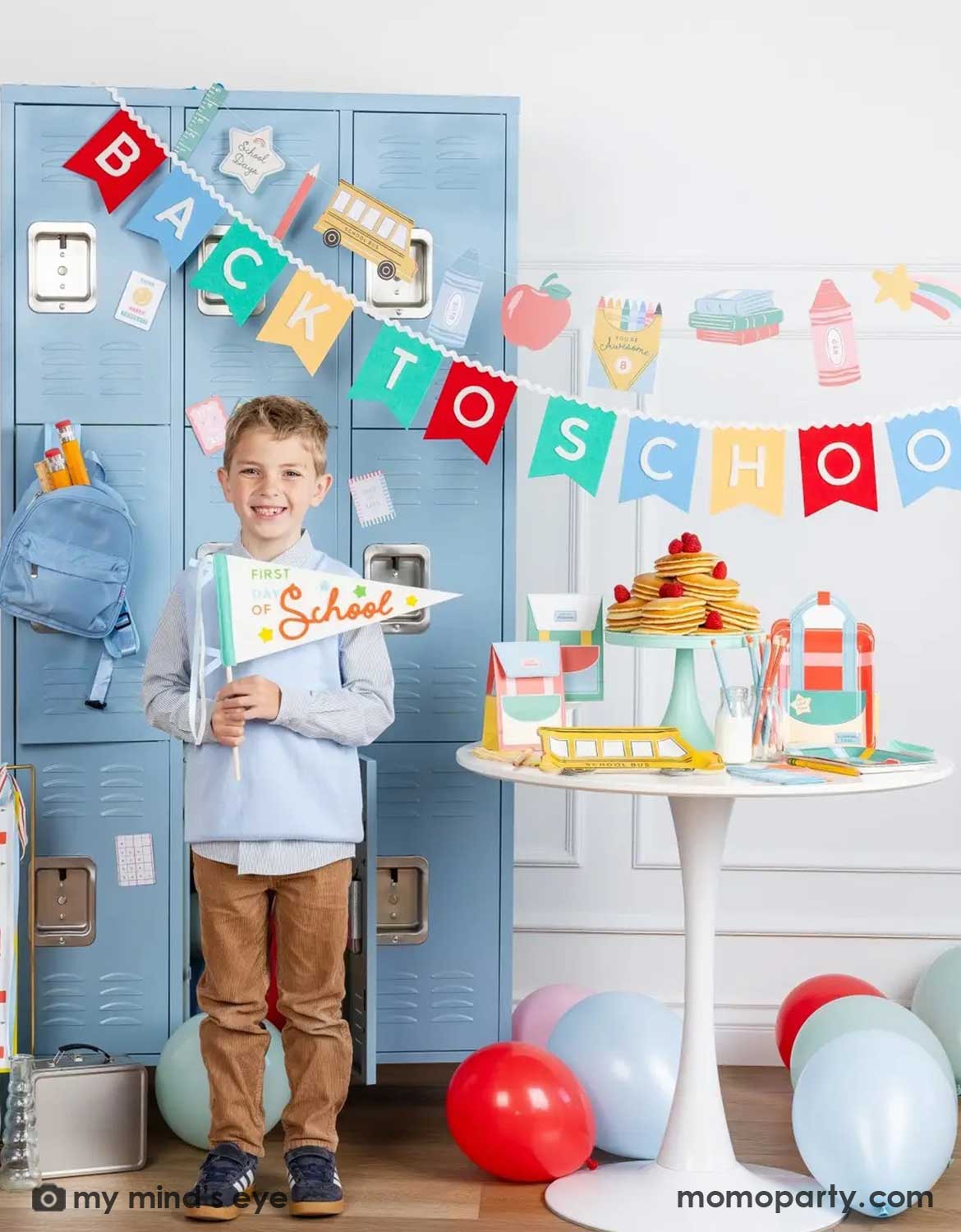 A boy holding the first day of school party pennant in a back to school party set up featuring various school themed party supplies and decors from Momo Party including school themed plates, treat boxes, party pennant, back to school felt garland along with school icons party banner hung above the light blue locker selves. A party table with pancakes on a cake stand next to some colorful backpack shaped treat bags. Along with colorful balloons spread across the room.